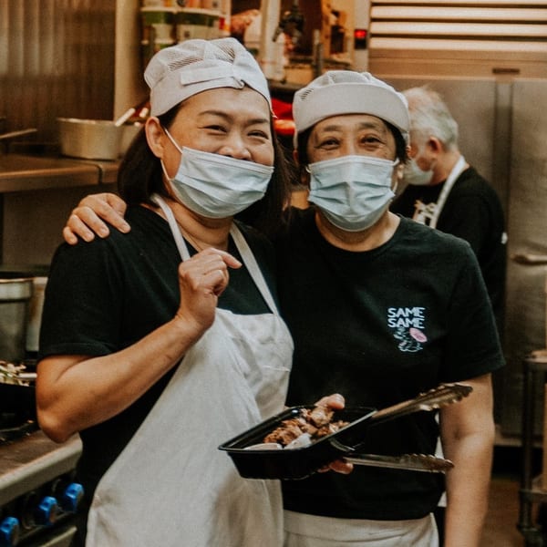 Two smiling Same Same Thai chefs in masks and aprons, one holding a to-go container of food.