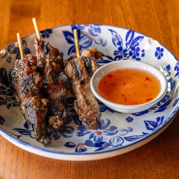 Grilled Thai pork skewers with a side of sweet chili dipping sauce on a blue and white plate.